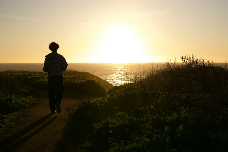 A person walking along a coastal path towards the setting sun