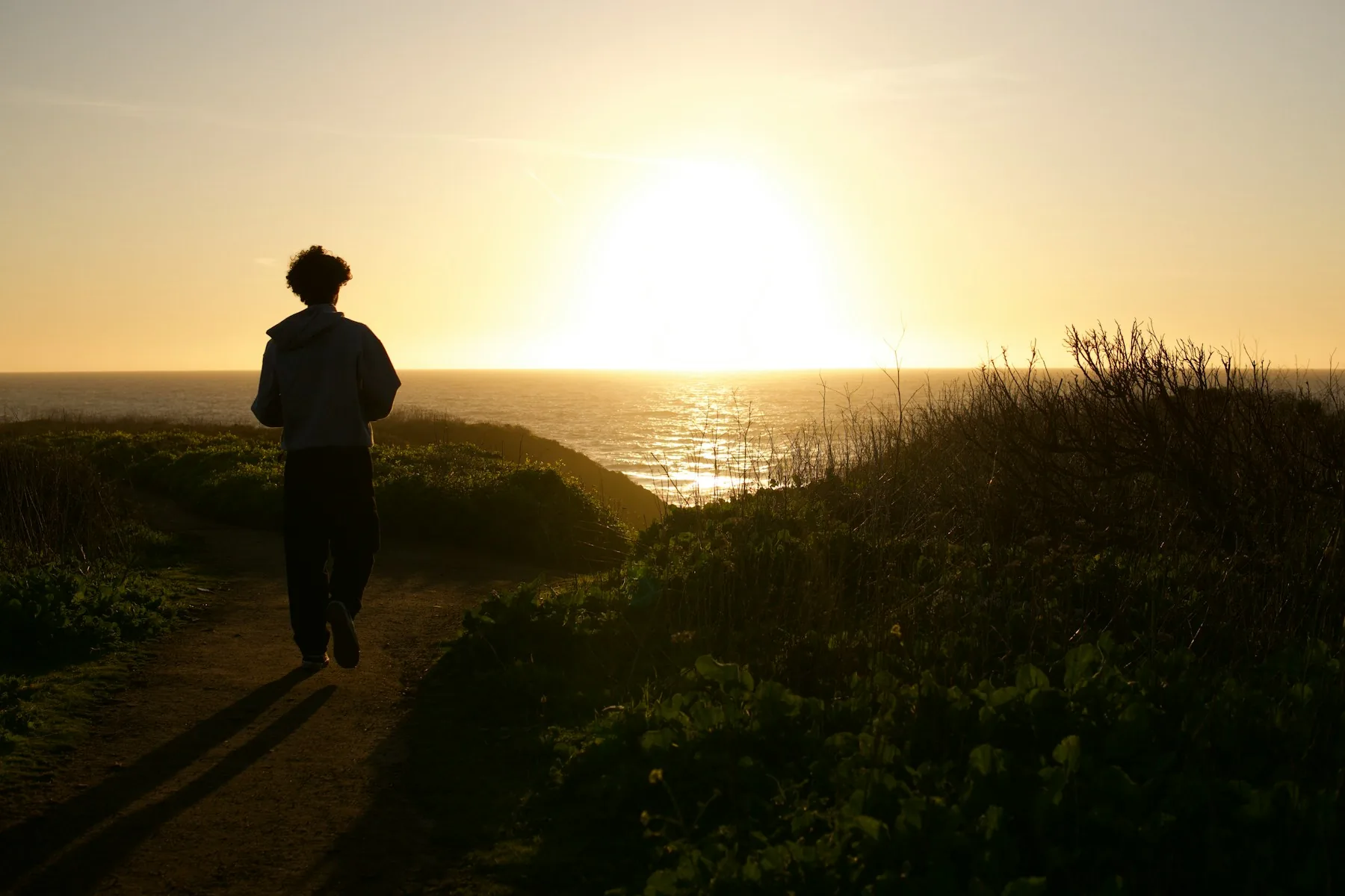 A person walking along a coastal path towards the setting sun