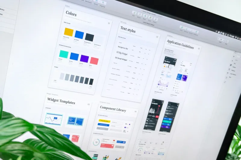 A monitor showing a design system with colour palettes, text styles and a component library, beside a leafy plant